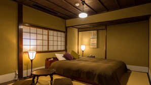 Un dormitorio de estilo japonés tradicional con suelo de tatami, una cama grande con muchas almohadas, una mesita y dos lámparas fijadas a la pared.