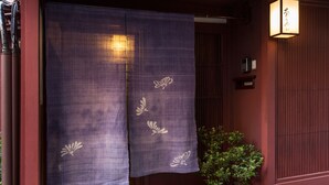 Entrada de una construcción japonesa tradicional con un fusuma morado, una planta en maceta y un pequeño estante con objetos.