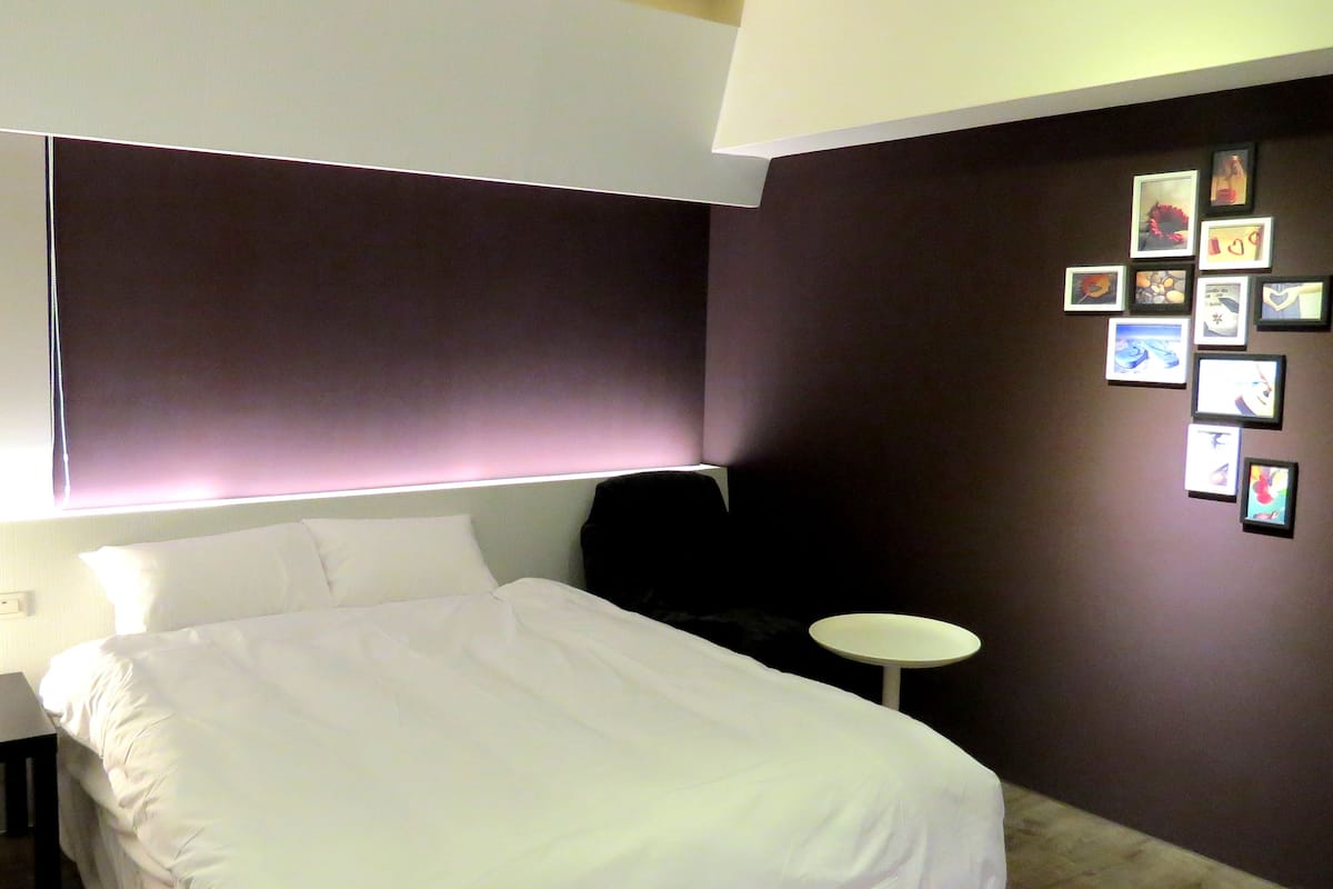 A hotel room with a bed, a small table, and a wall with framed pictures.
