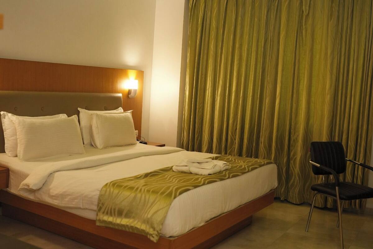 Una habitación de hotel con cama, almohadas, una lámpara de noche, una silla y una cortina.