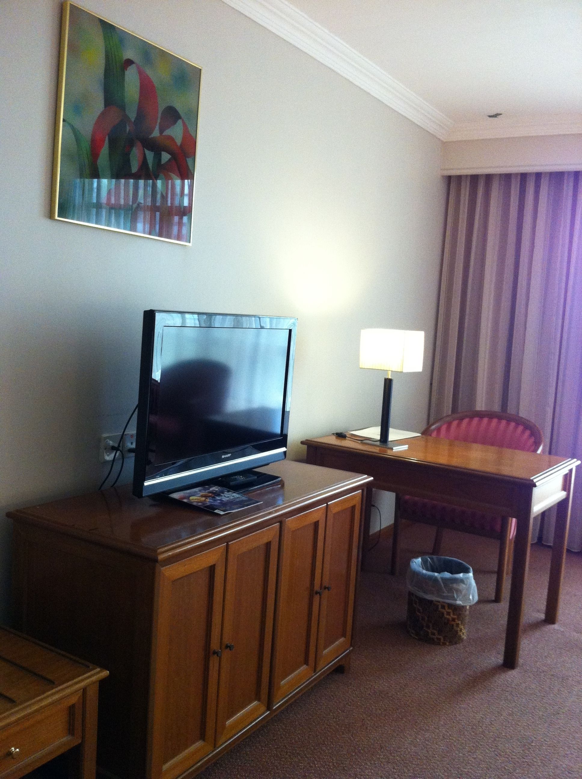 A hotel room with a flat-screen TV on a wooden cabinet, a small table with a lamp, a chair, and a trash can.