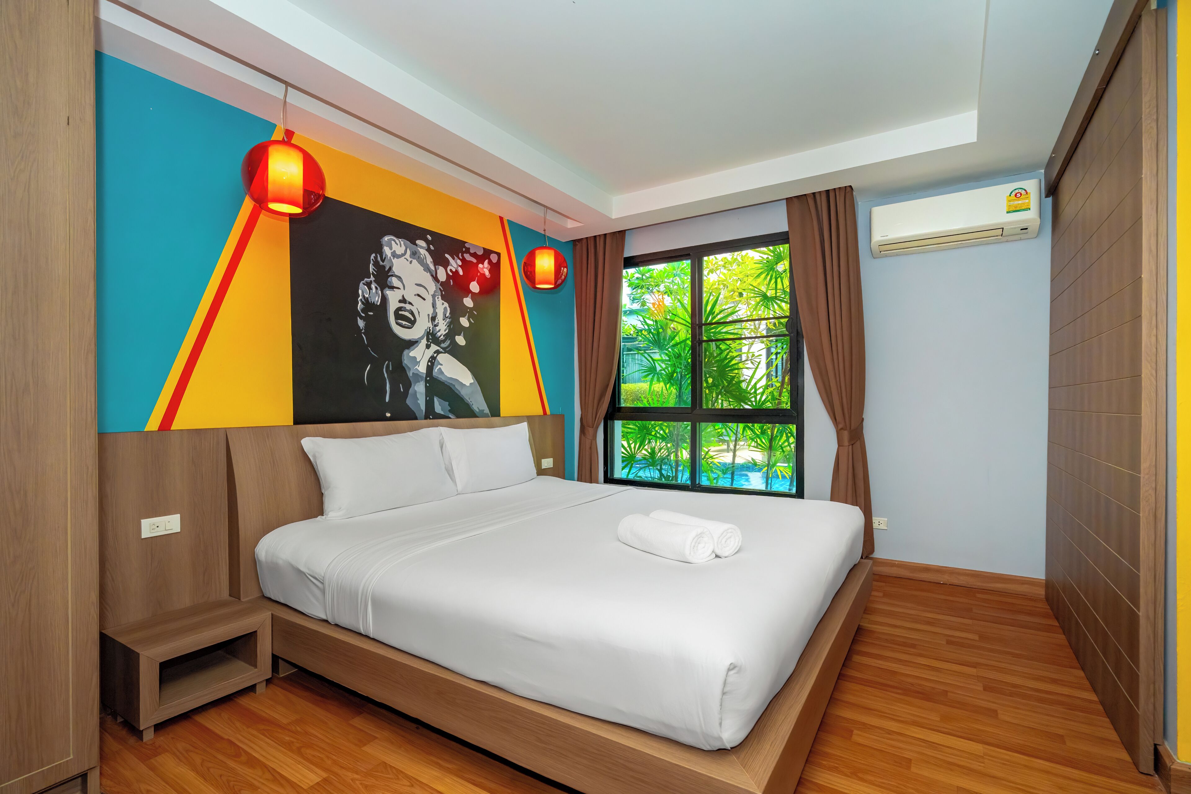 Una habitación de hotel moderna con una cama grande, un mural colorido, una ventana con vistas a la vegetación y un equipo de aire acondicionado.