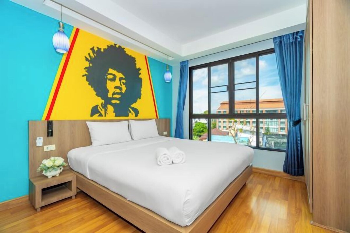 Habitación de hotel con una cama grande, un cabecero con un cuadro, una mesita de noche con un jarrón y una ventana con cortinas.