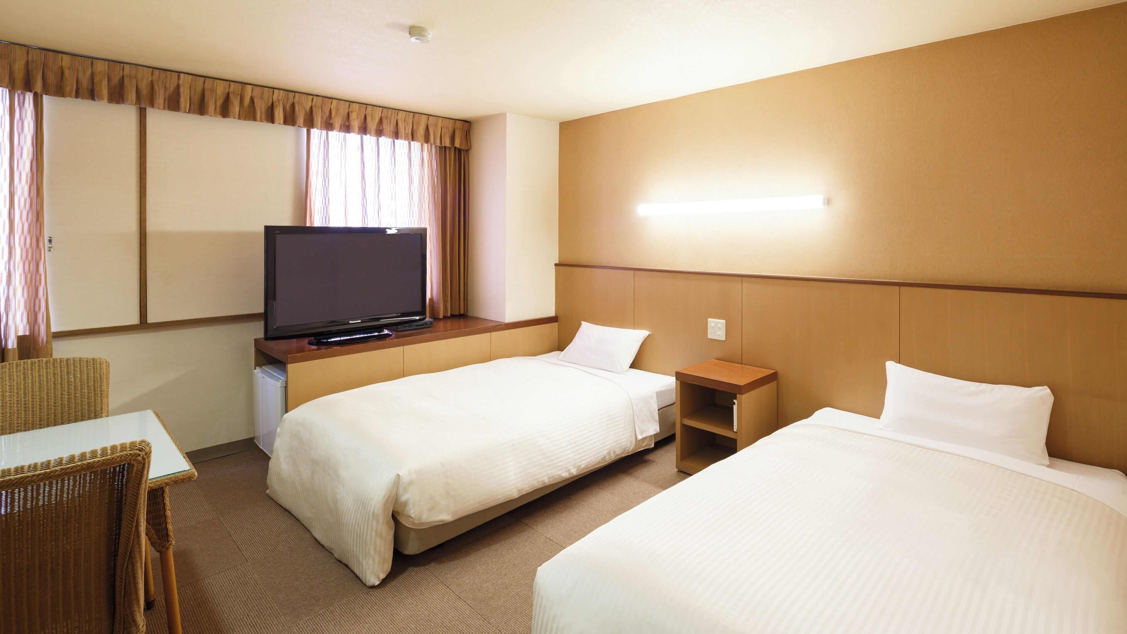 Quarto de hotel com duas camas, televisão sobre um suporte, uma mesinha e uma cadeira.