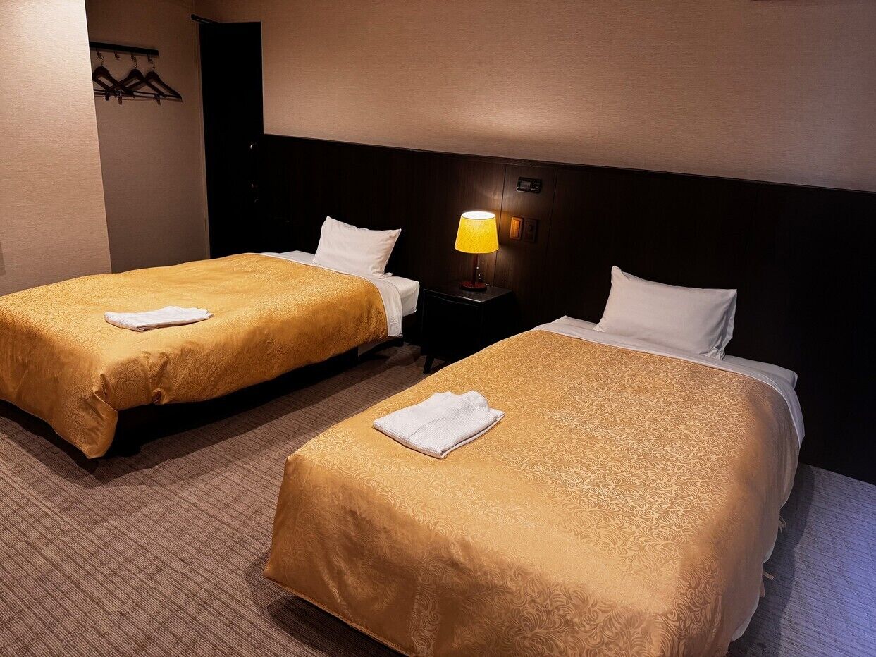 Habitación de hotel con dos camas, una mesita de noche con lámpara y un perchero.