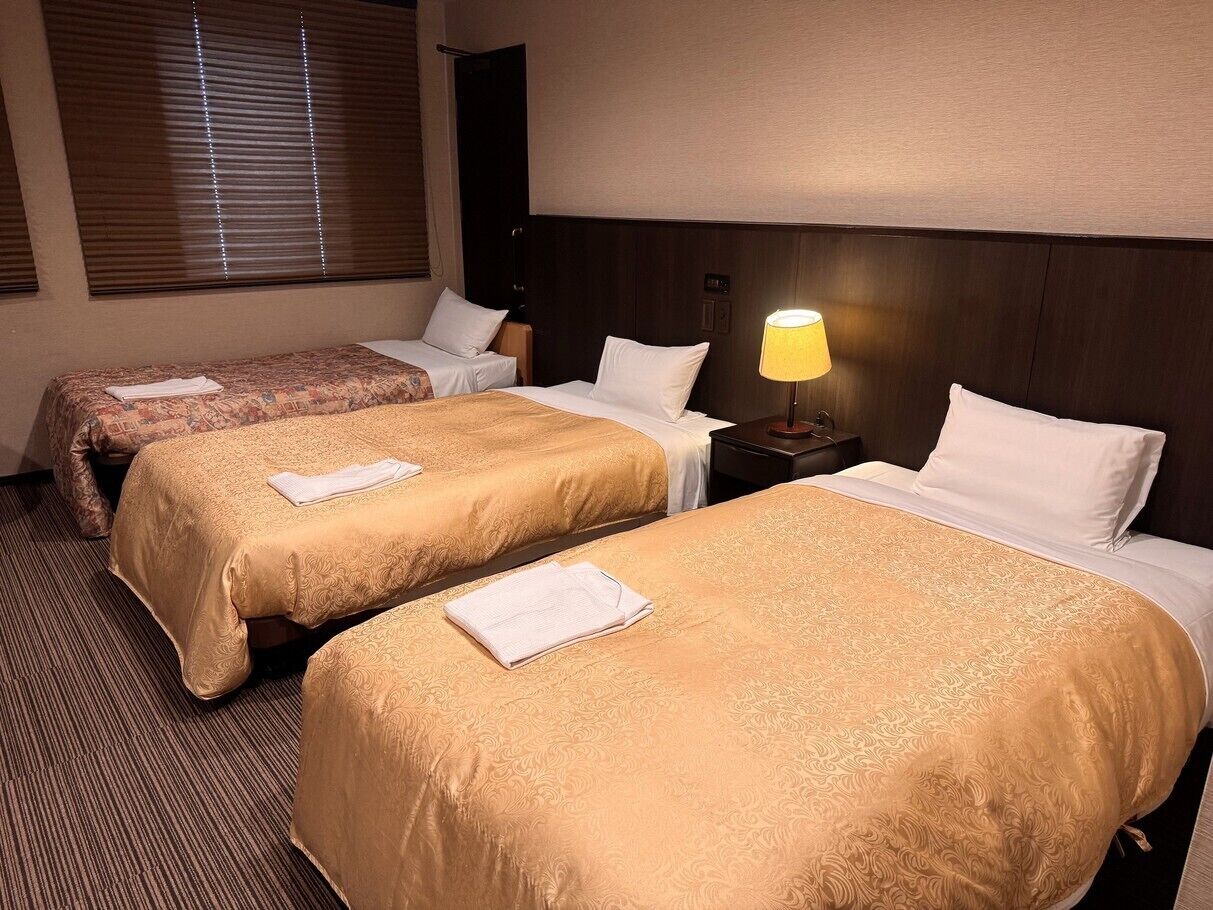 Una habitación de hotel con dos camas, una mesita de noche, una lámpara y un ventanal con persianas.