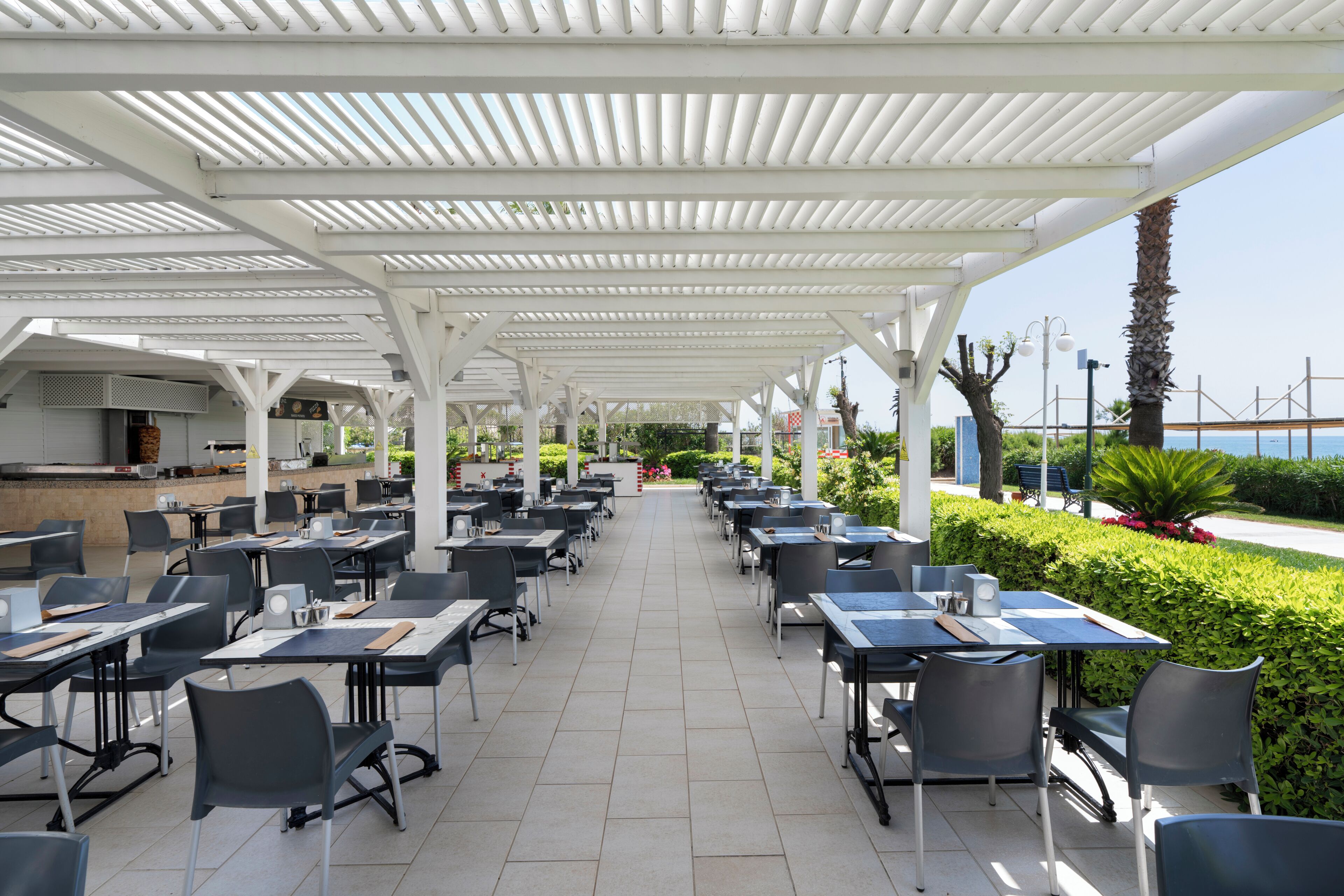 A covered outdoor dining area with tables and chairs, a grill station, and a view of the sea.