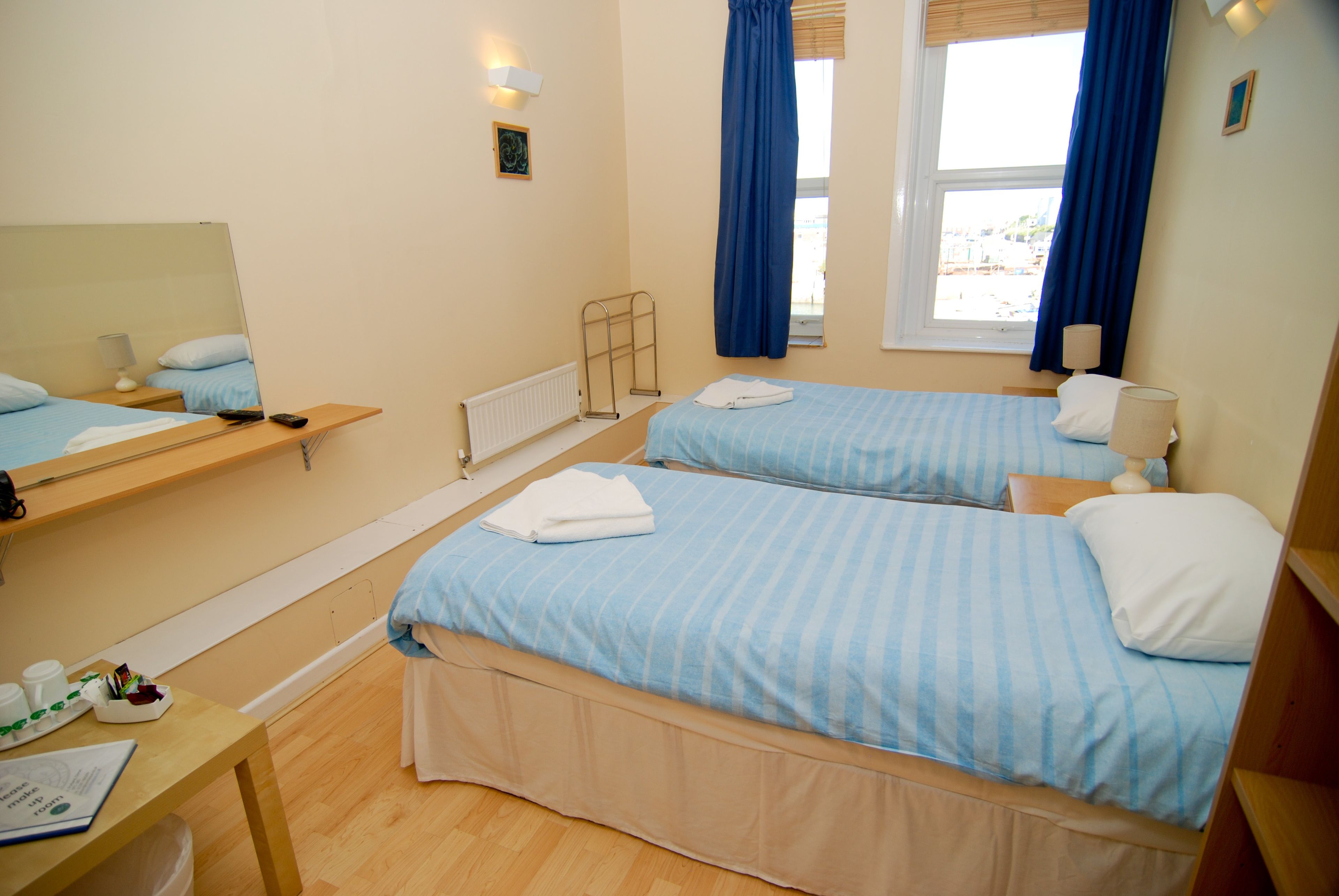A hotel room with two beds, a wooden table, a mirror, a window with curtains, and a small picture on the wall.