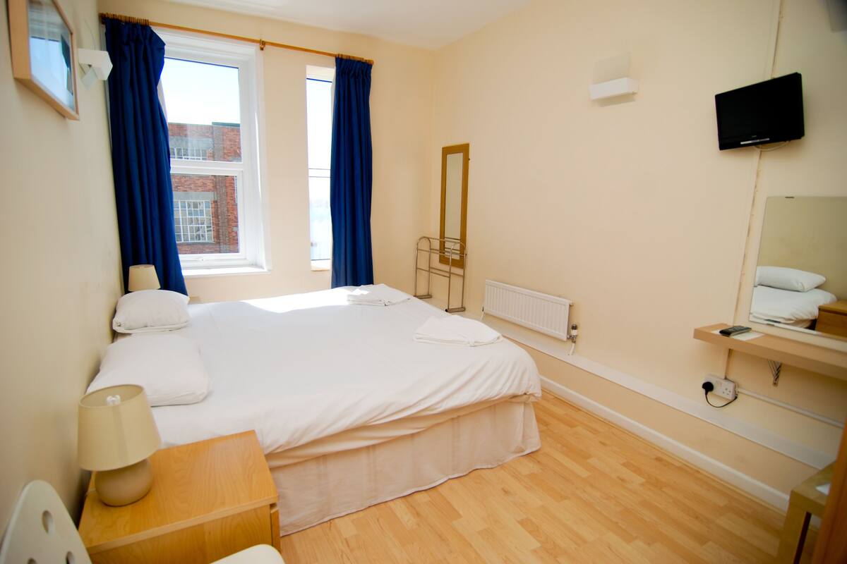 Quarto de hotel com uma cama, mesa de cabeceira com abajur, televisão na parede e janela com cortinas.