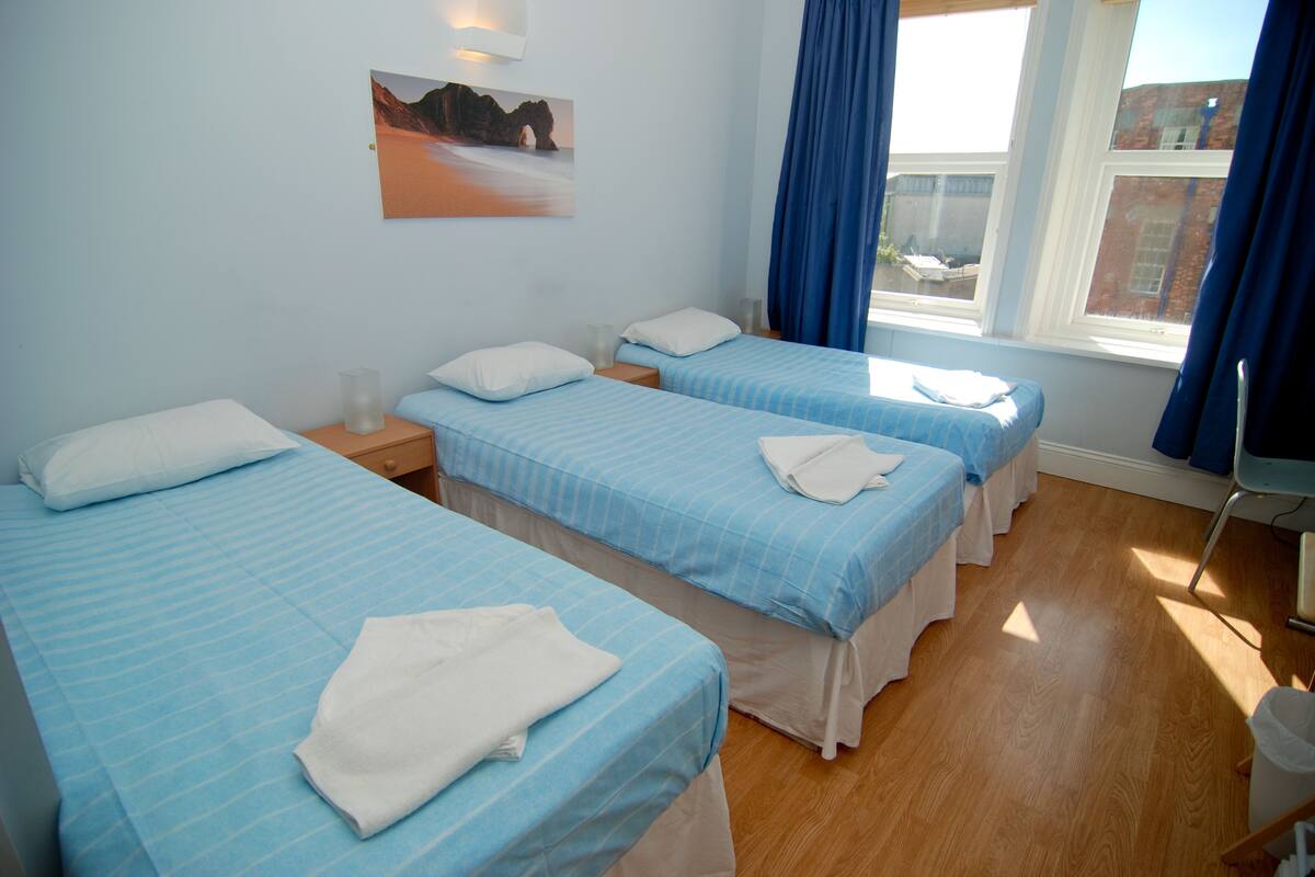 Quarto de hotel com duas camas, um quadro na parede, uma janela com cortinas e piso de madeira.