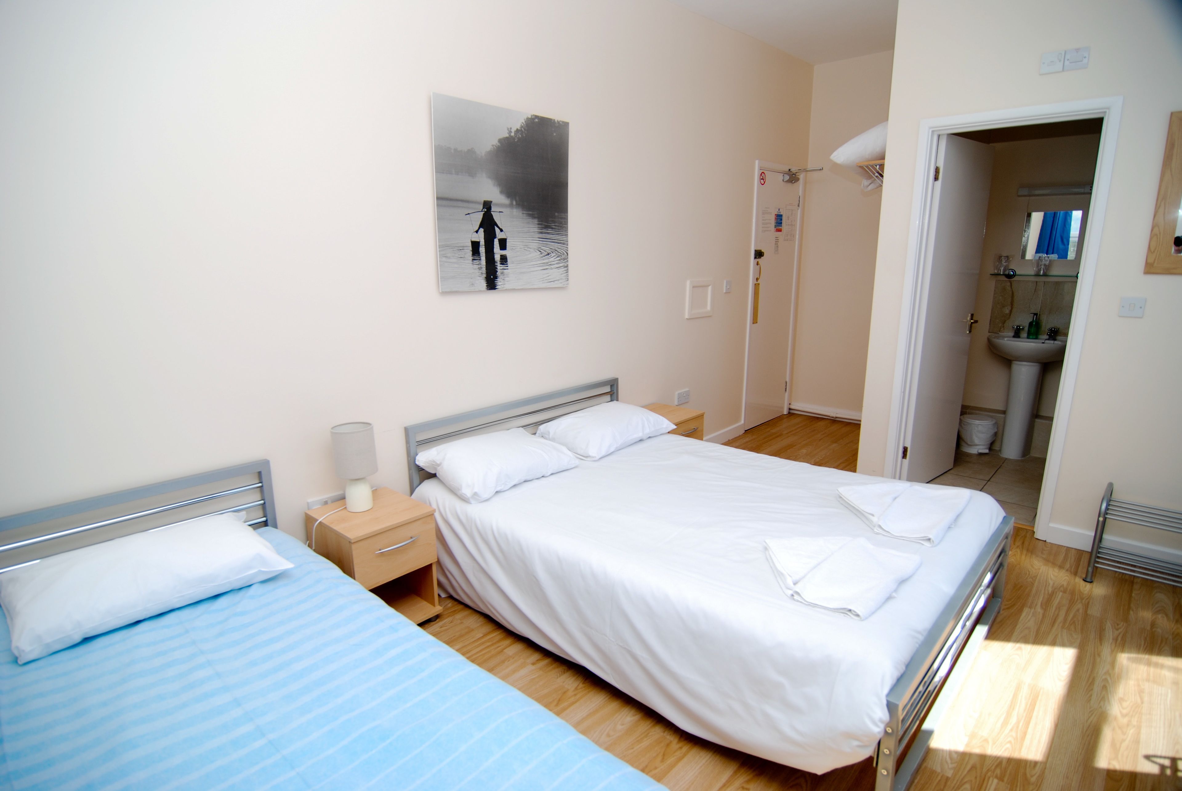 A hotel room with a large bed, a smaller bed, a nightstand, a lamp, a painting on the wall, and a bathroom visible through an open door.