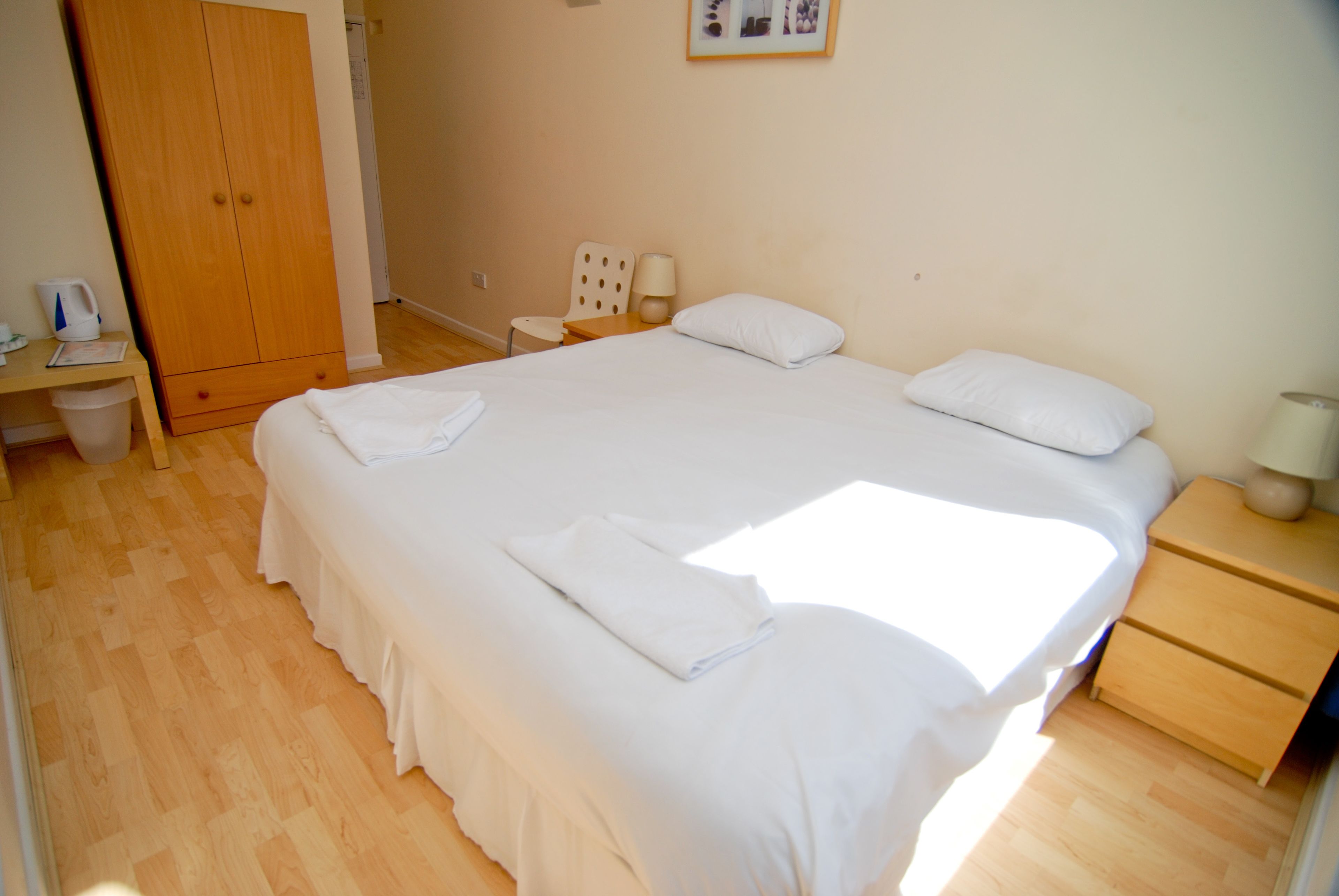 A bedroom with a large bed, a wooden wardrobe, a small table with a chair, and a bedside table with a lamp.