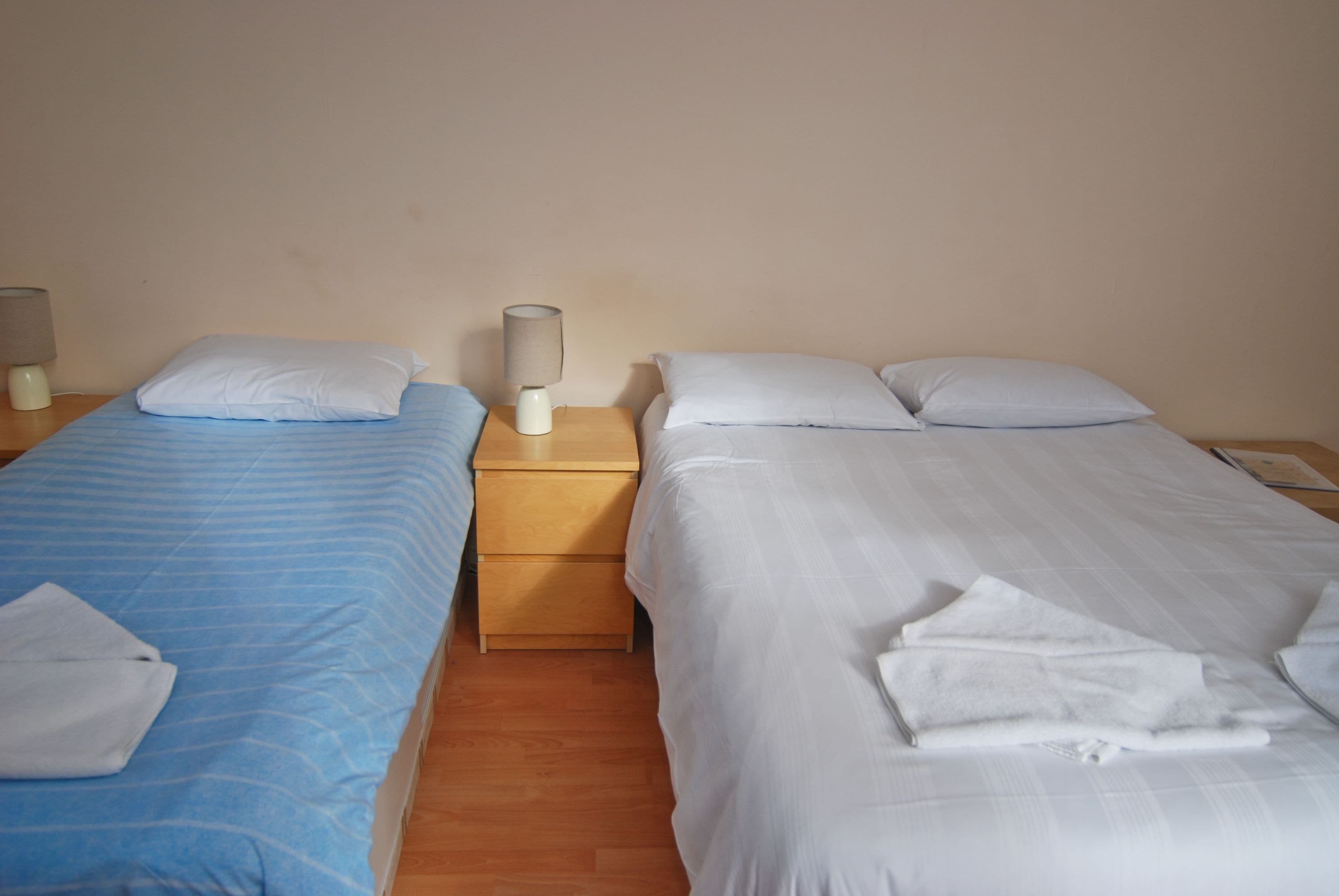 A hotel room with two beds, each with a pillow and a folded towel. There is a nightstand with a lamp between the beds.