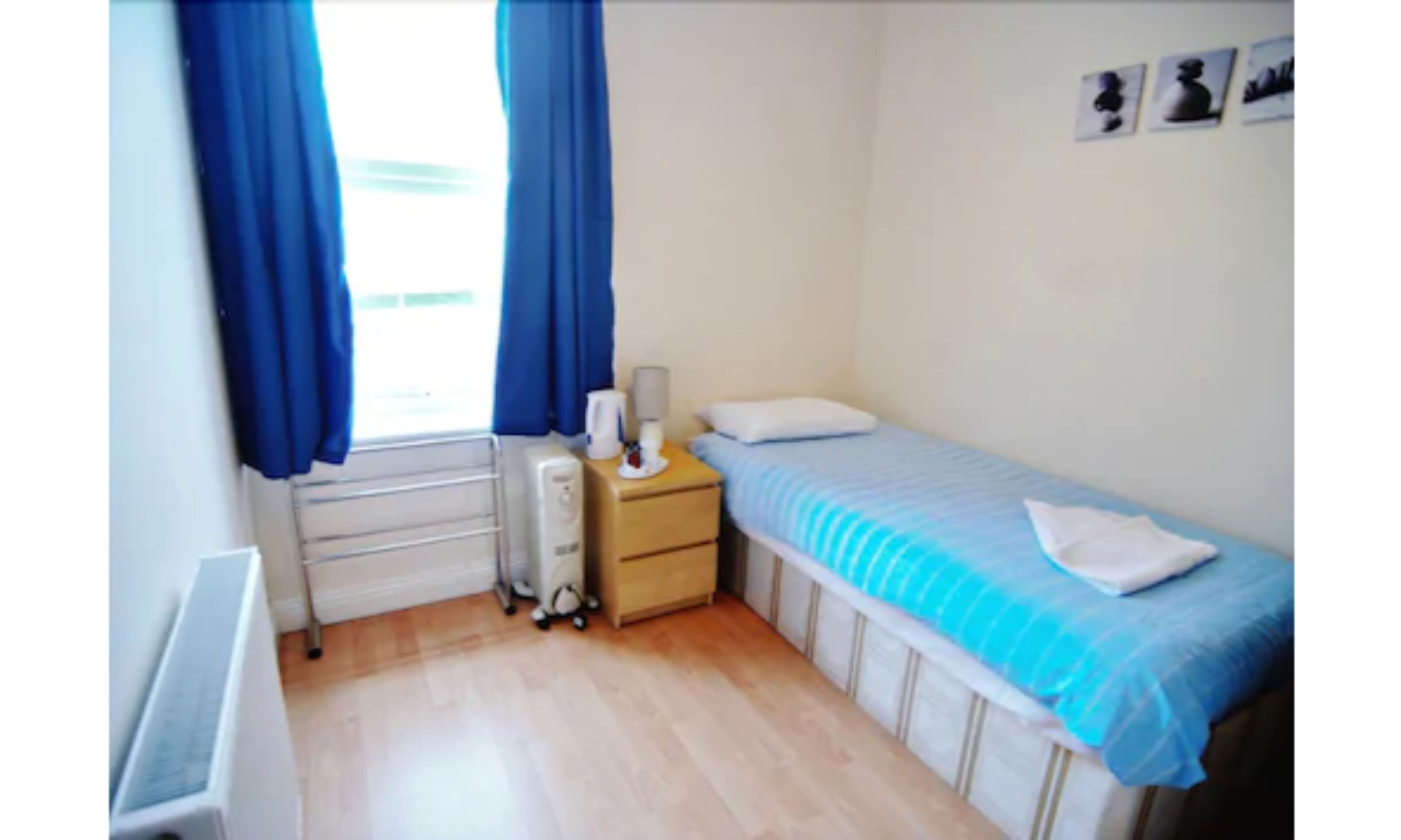 Ein kleines, ordentliches Zimmer mit einem Einzelbett, einem Nachttisch mit Lampe, einem Heizkörper und einem Fenster mit Vorhängen.