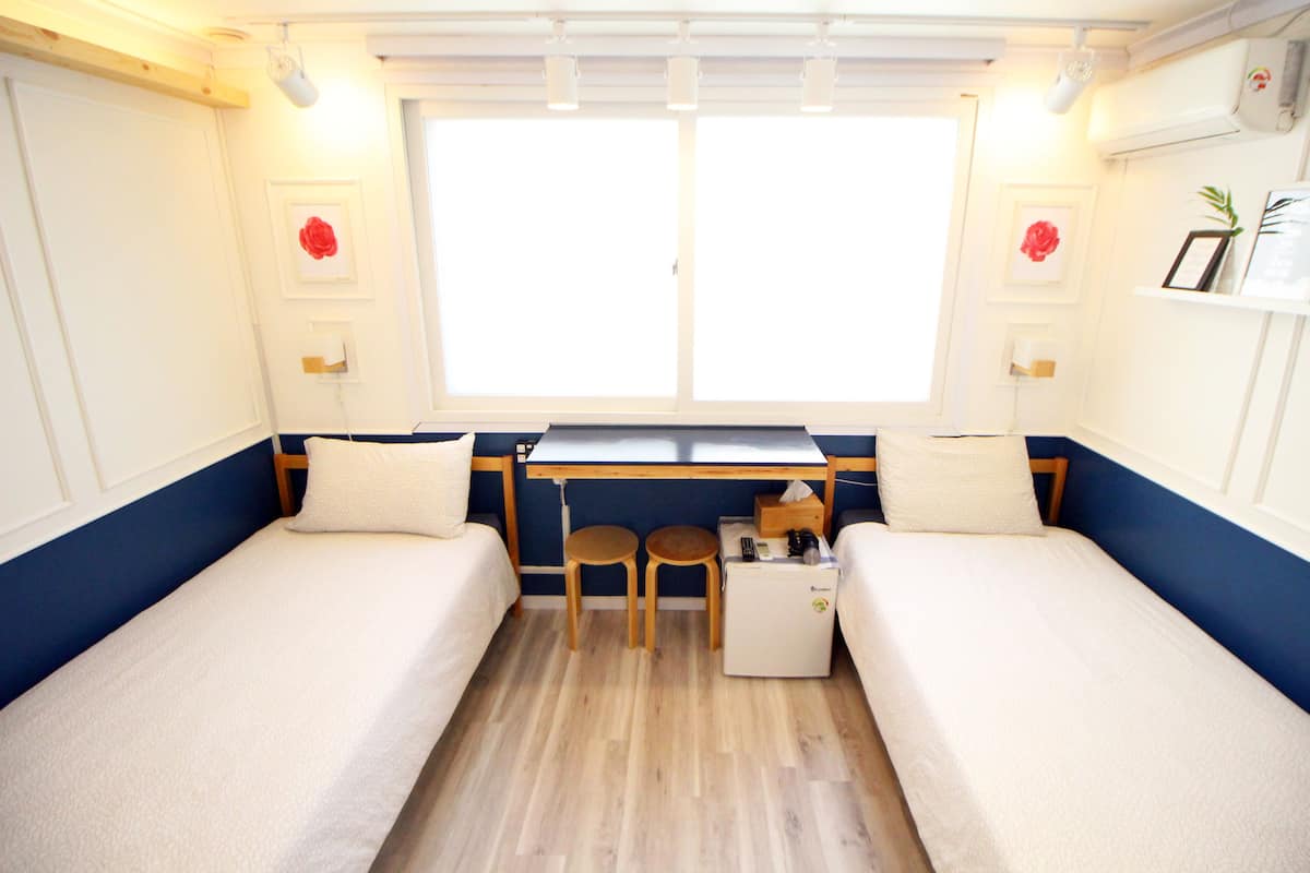 Une chambre de taille modeste, équipée de deux lits simples, d’une petite table, de deux chaises et d’un coin cuisine compact.
