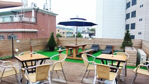 Une terrasse extérieure avec des tables en bois et des chaises en osier, un barbecue et une vue sur les bâtiments en arrière-plan.