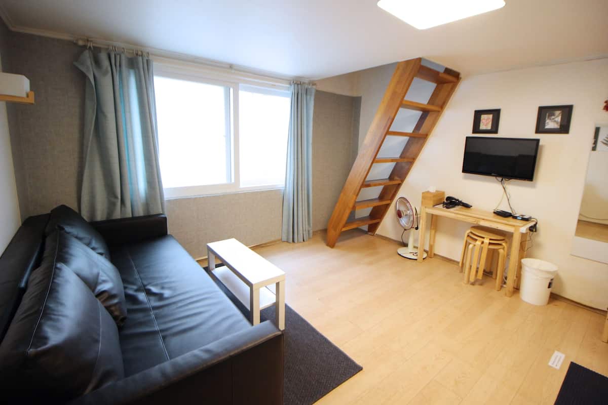 Un espace de vie compact comprenant un canapé en cuir noir, une petite table blanche, un bureau en bois avec une chaise, un téléviseur fixé au mur et une échelle en bois.