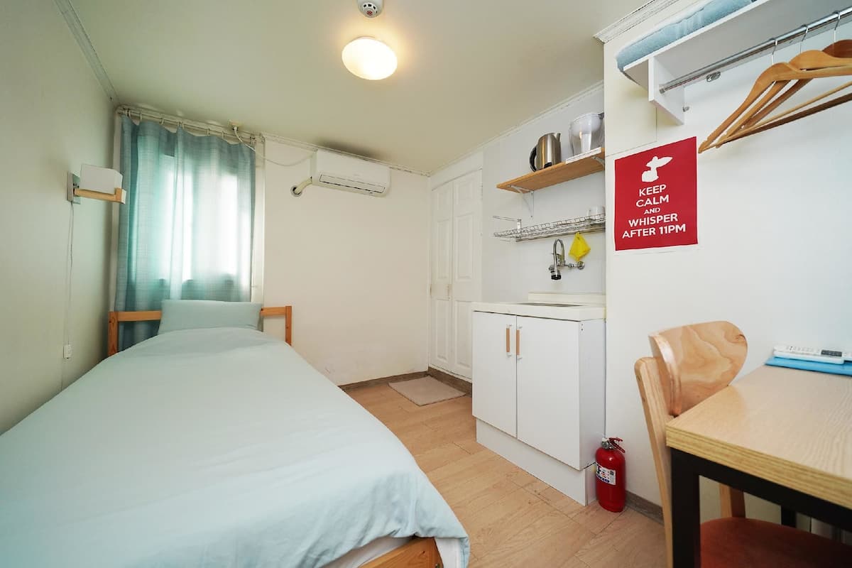 Une chambre de taille modeste comprenant un lit, une petite kitchenette, une table à manger et un panneau indiquant qu’il faut rester calme et parler à voix basse après 23 heures.