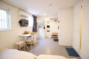 A compact hotel room with a bed, a desk with a chair, a TV mounted on the wall, a small kitchenette, and a bathroom visible through an open door.