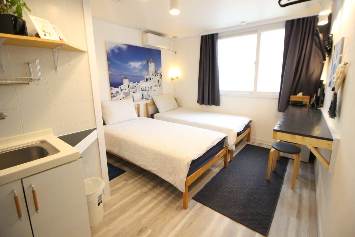 Une chambre d’hôtel compacte comprenant deux lits simples, une petite kitchenette, un bureau et une grande fenêtre avec des rideaux.
