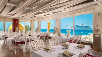 Un restaurant avec des tables nappées de blanc, des coussins rouges et une vue sur la mer.
