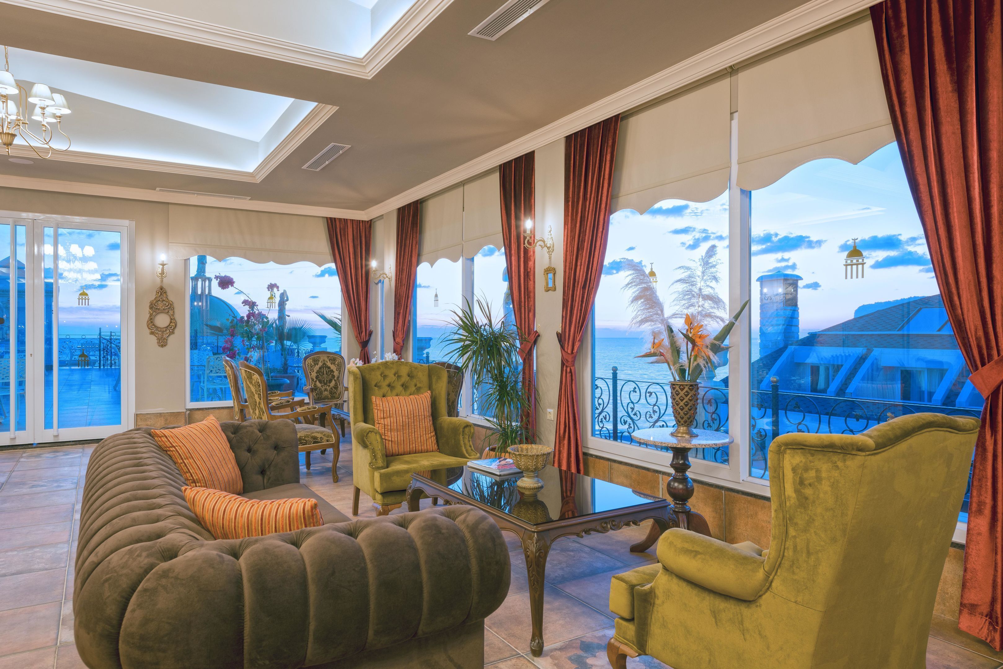 Un salón amplio con un sofá grande, sillones y una mesa de centro. Cuenta con ventanas grandes con cortinas, que ofrecen vistas al mar y un edificio.