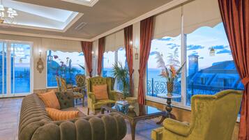 Un salón amplio con un sofá grande, sillones y una mesa de centro. Cuenta con ventanas grandes con cortinas, que ofrecen vistas al mar y un edificio.