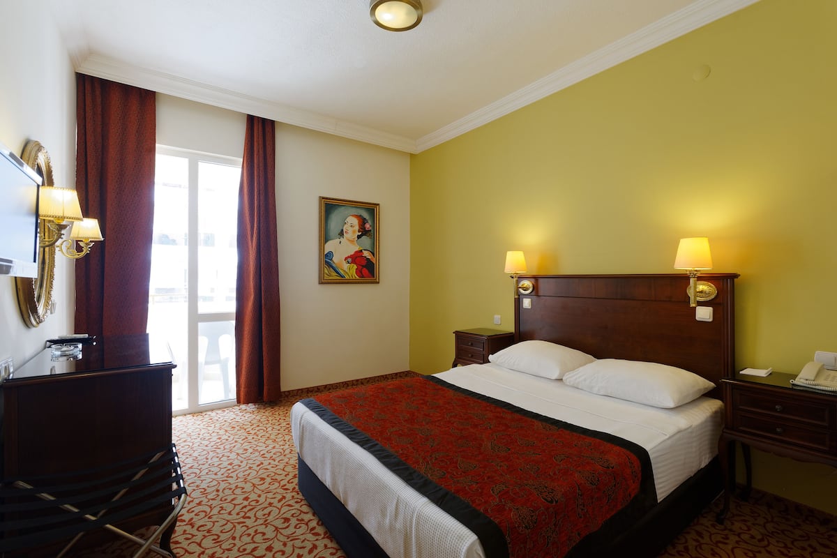 A hotel room with a large bed, a desk, a chair, a desk lamp, a nightstand, a telephone, and a painting on the wall.