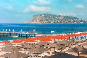 A beach with rows of umbrellas and lounge chairs, a pier extending into the sea, and a hill with buildings in the background. - Antique Roman Palace Museum Hotel - Adults Only Ultra All Inclusive (Alanya)