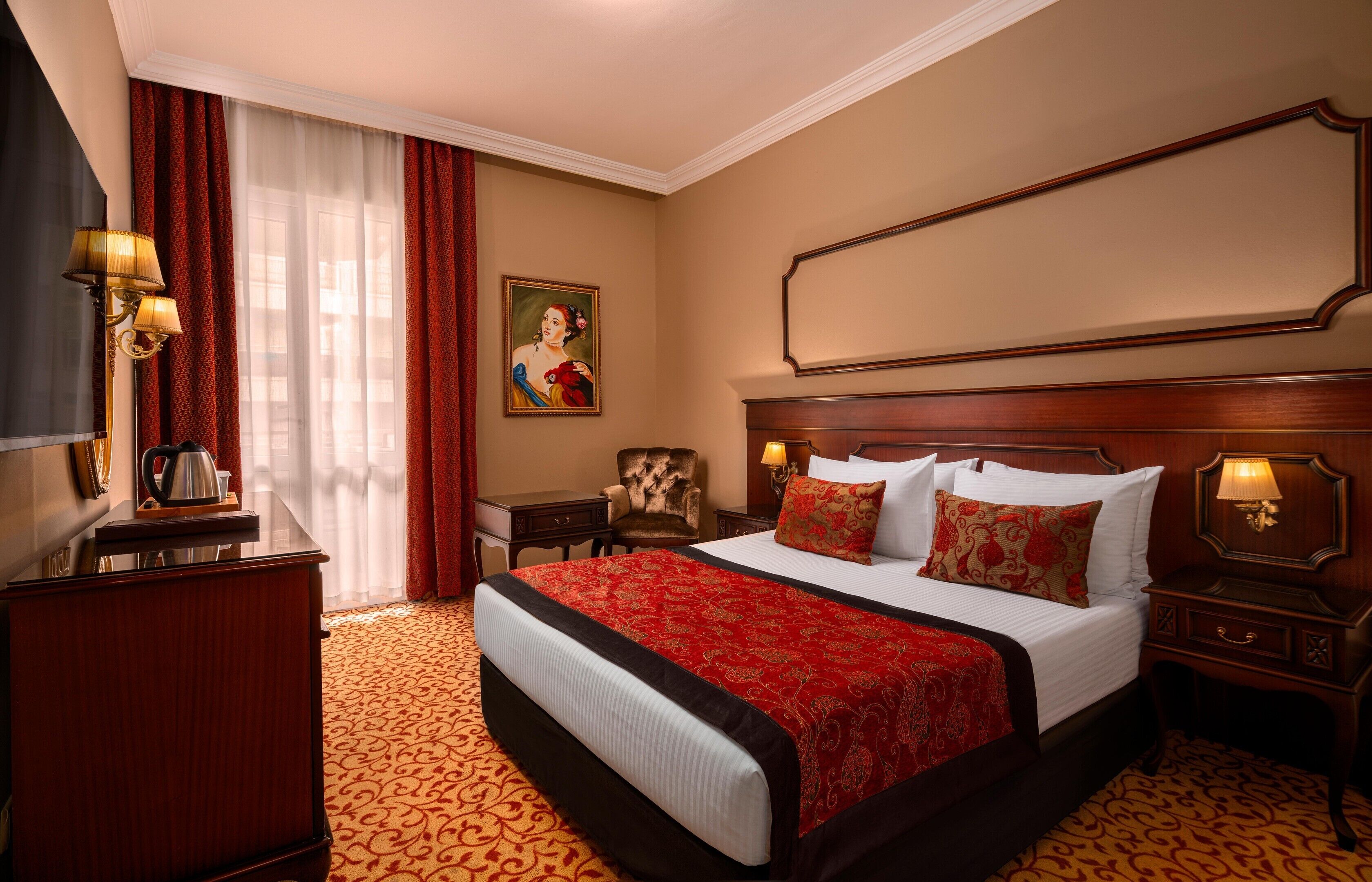 Habitación de hotel con una cama grande, un escritorio, una silla, un cuadro en la pared y un ventanal con cortinas.