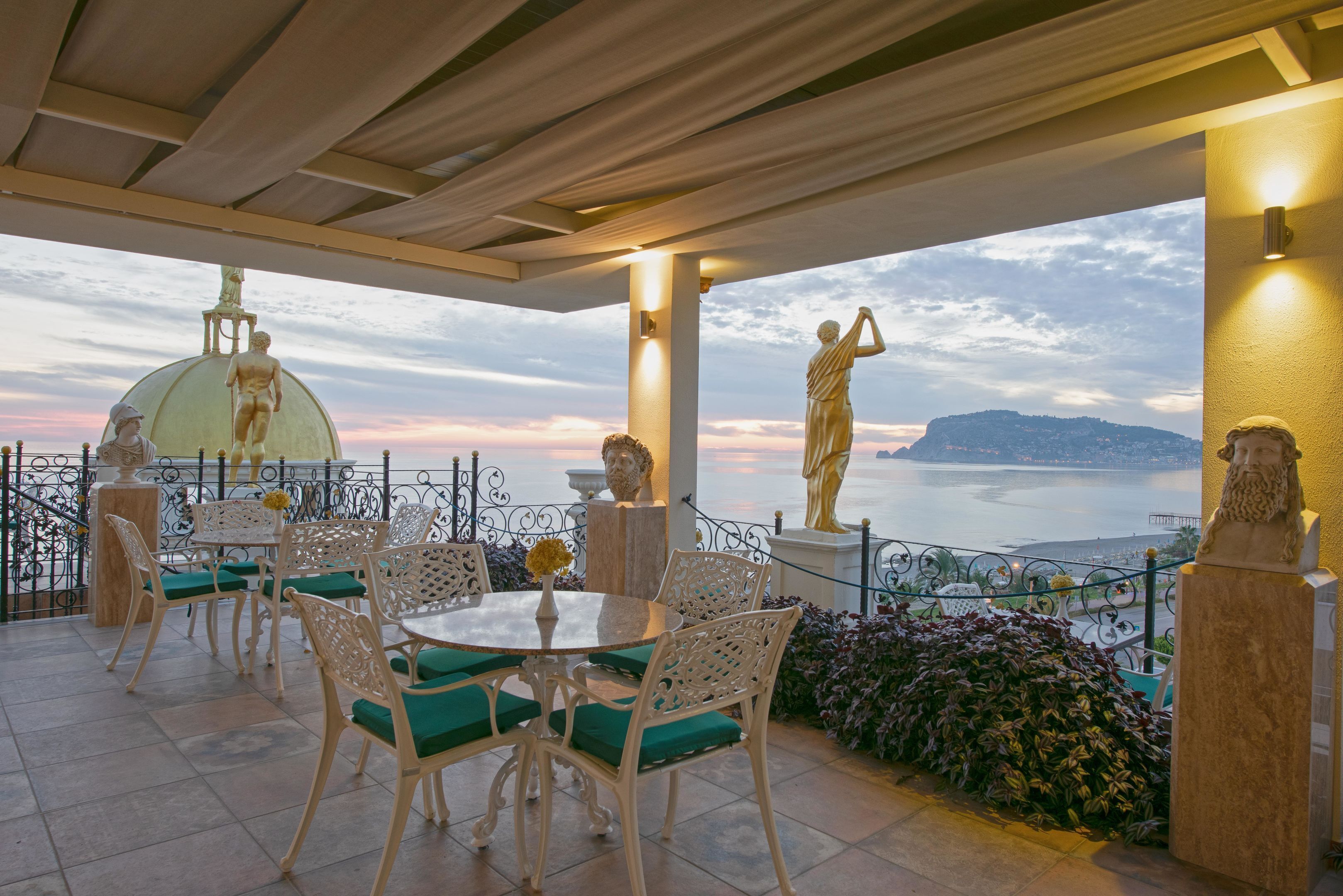 Un patio al aire libre con mobiliario de mimbre blanco, una mesa redonda y cojines verdes. Se observan estatuas y una vista al mar.