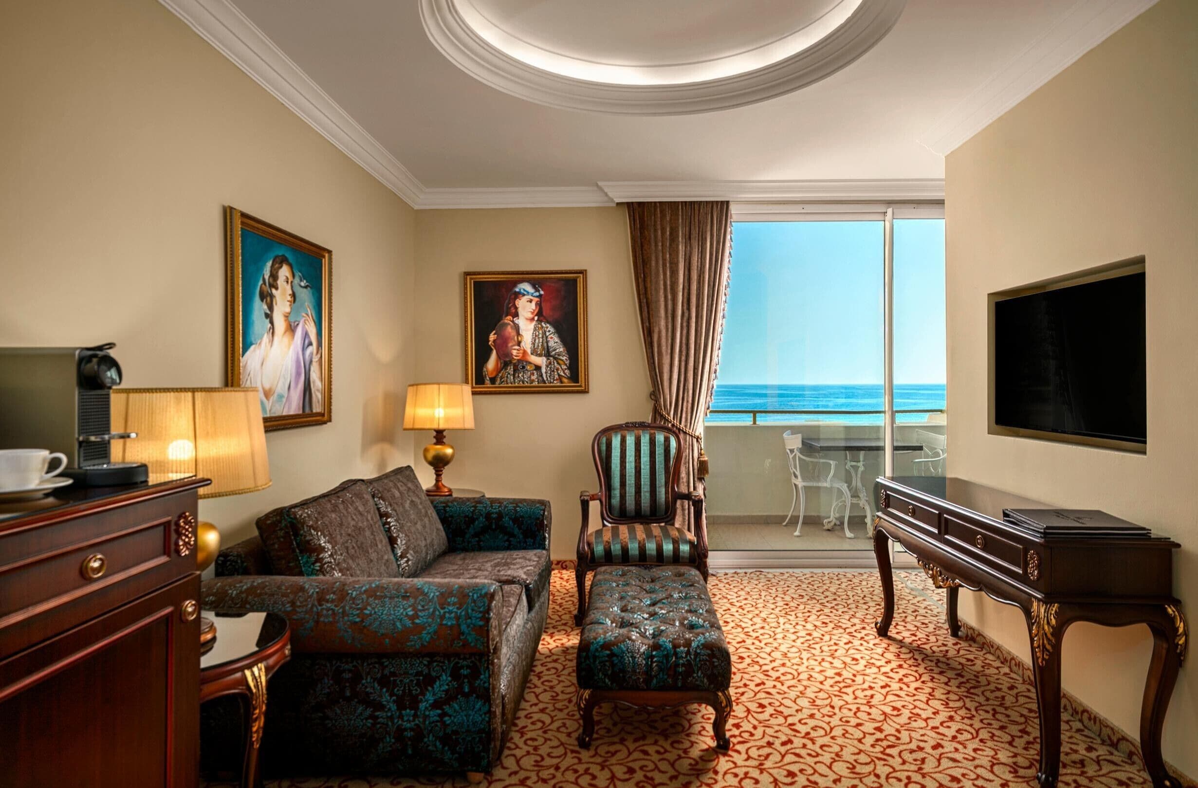 A hotel room with a sofa, armchair, and a desk with a flat-screen TV. There is a painting on the wall and a view of the sea through the window.