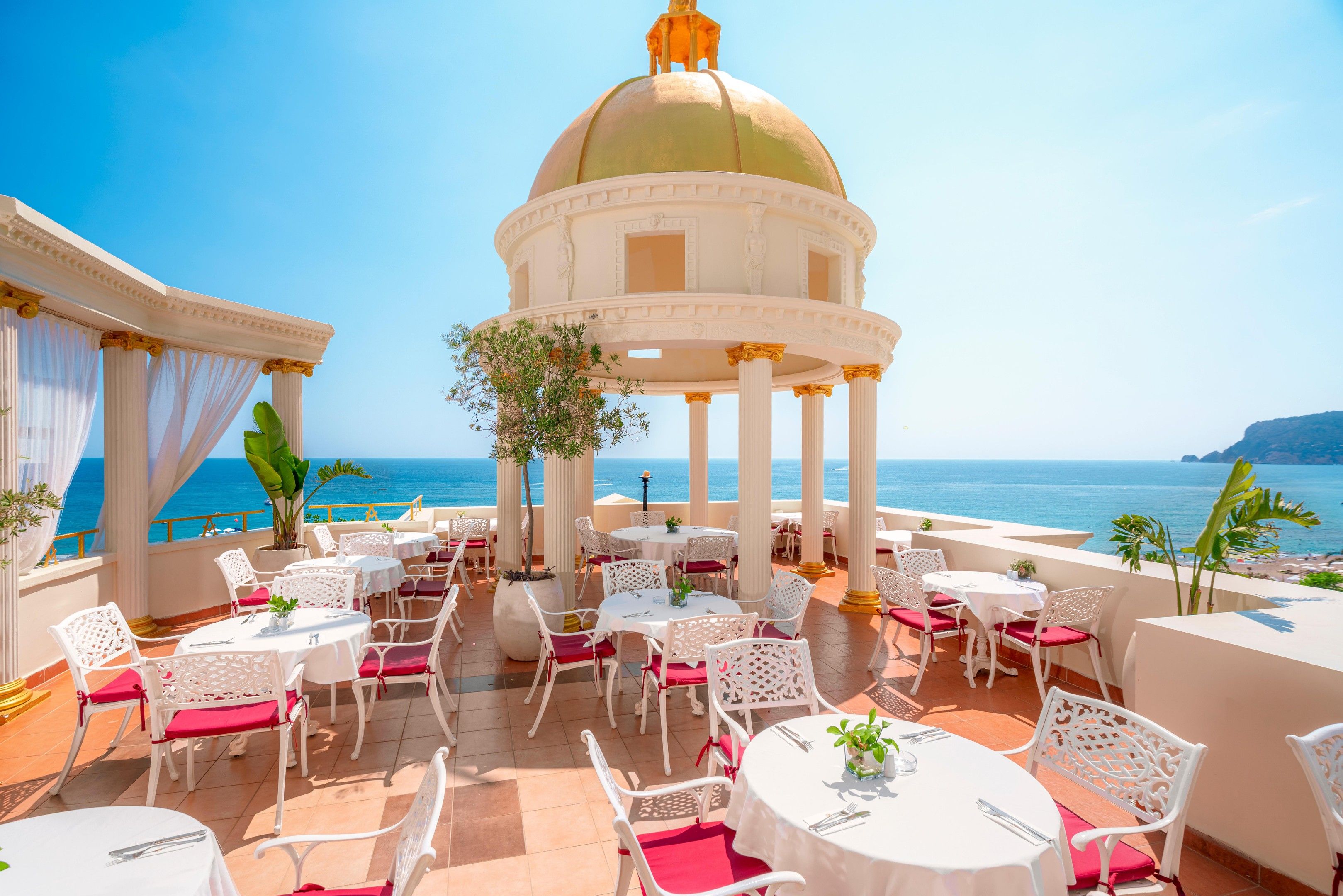 An outdoor dining area with white tables and chairs, a central columned structure, and a view of the sea.