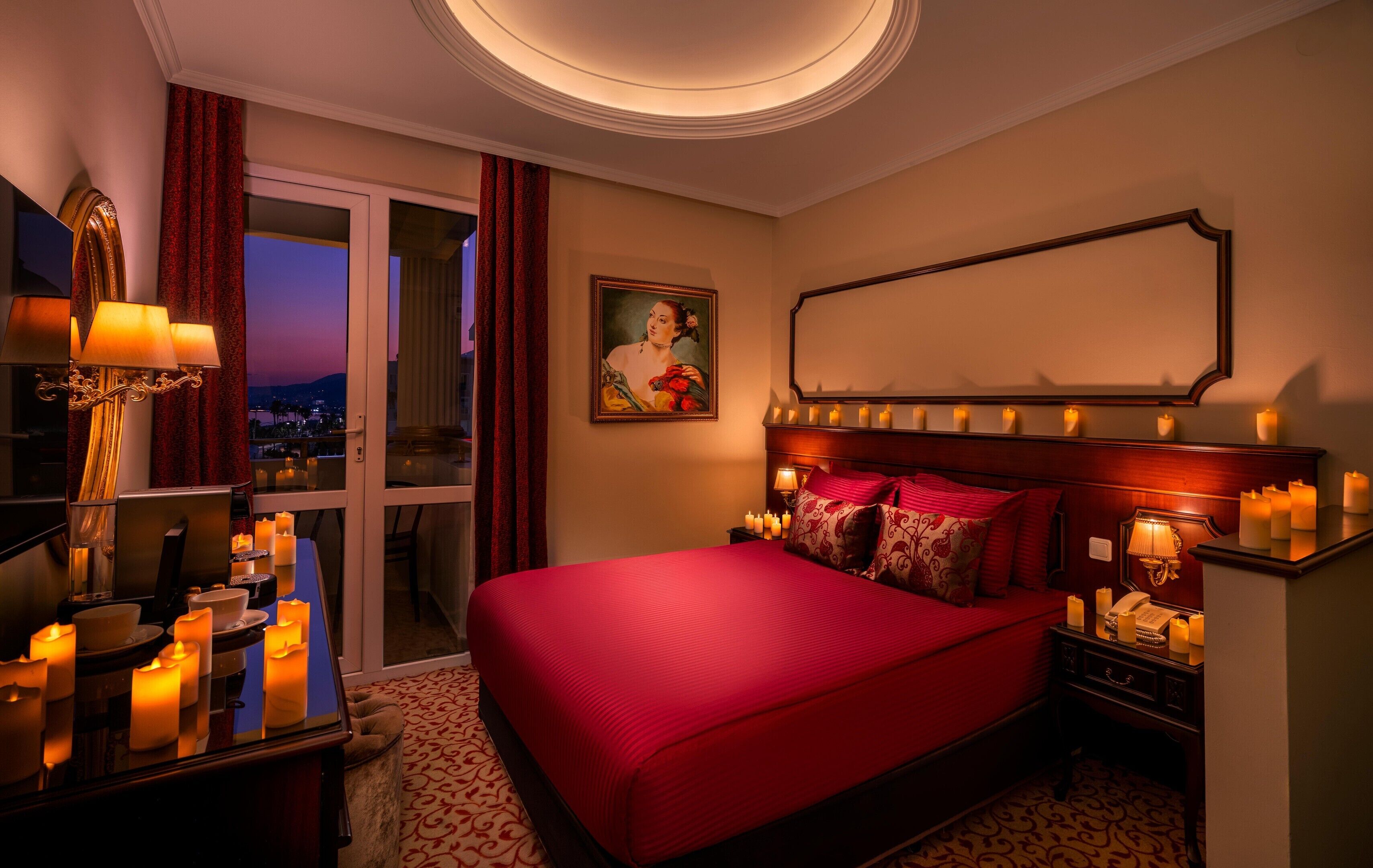 A hotel room with a large bed, a painting on the wall, a mirror above the bed, and multiple candles on the nightstand.