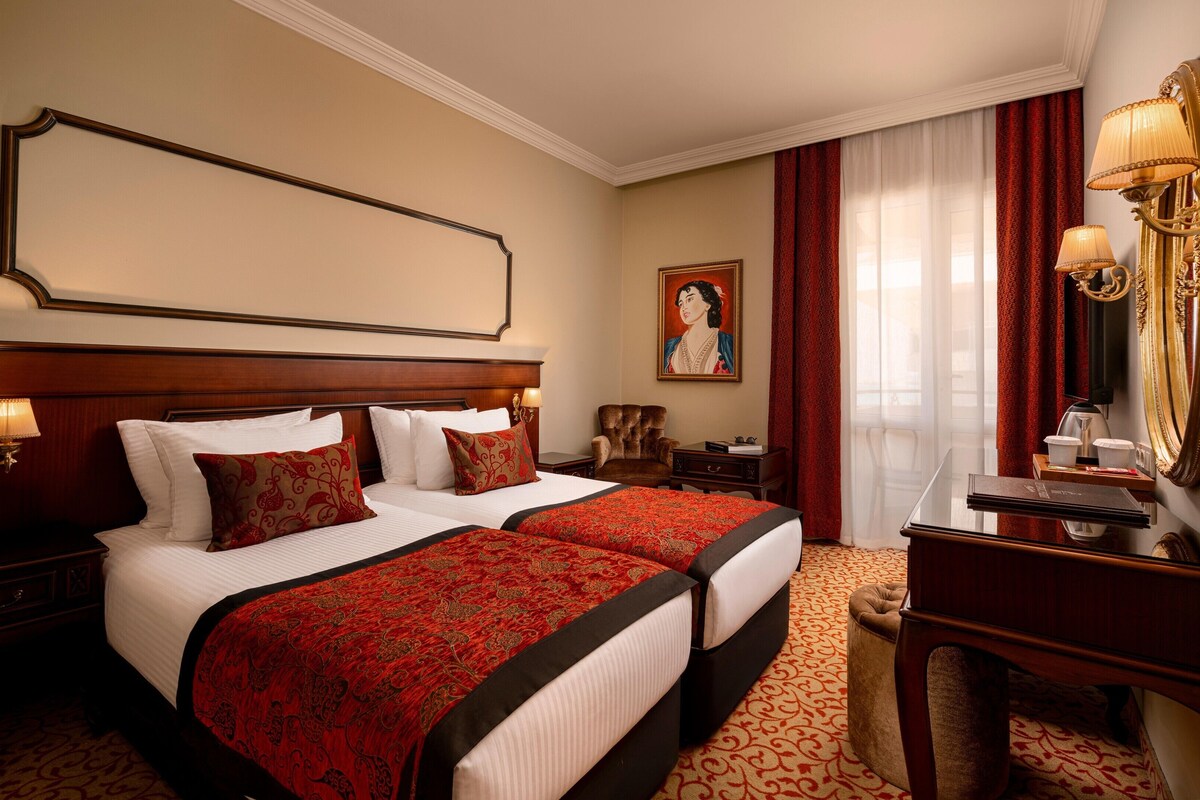 A hotel room with two beds, a large mirror, a desk with a lamp, and a painting on the wall.