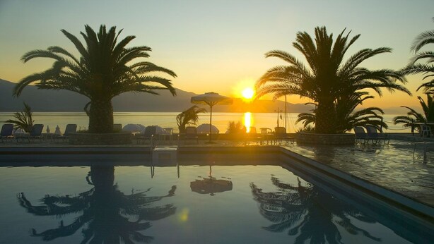 Un espace aménagé au bord de la piscine, avec des chaises longues et des palmiers, où se reflète le coucher de soleil sur l’eau.