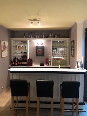 Een bar met een wit aanrecht, drie zwarte barkrukken en planken vol flessen en glazen.