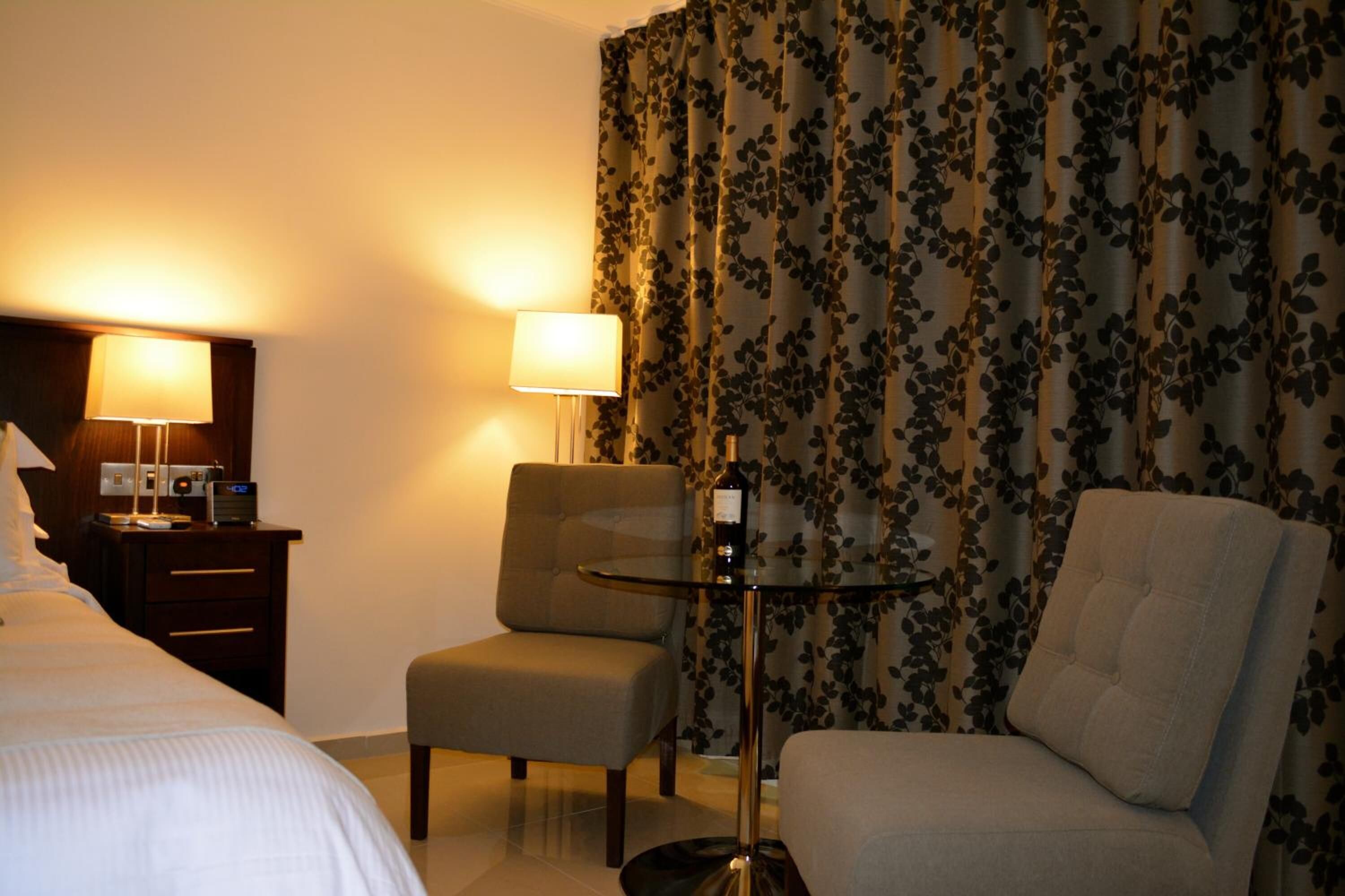 A hotel room with a bed, two armchairs, a small table with a bottle, and a lamp.