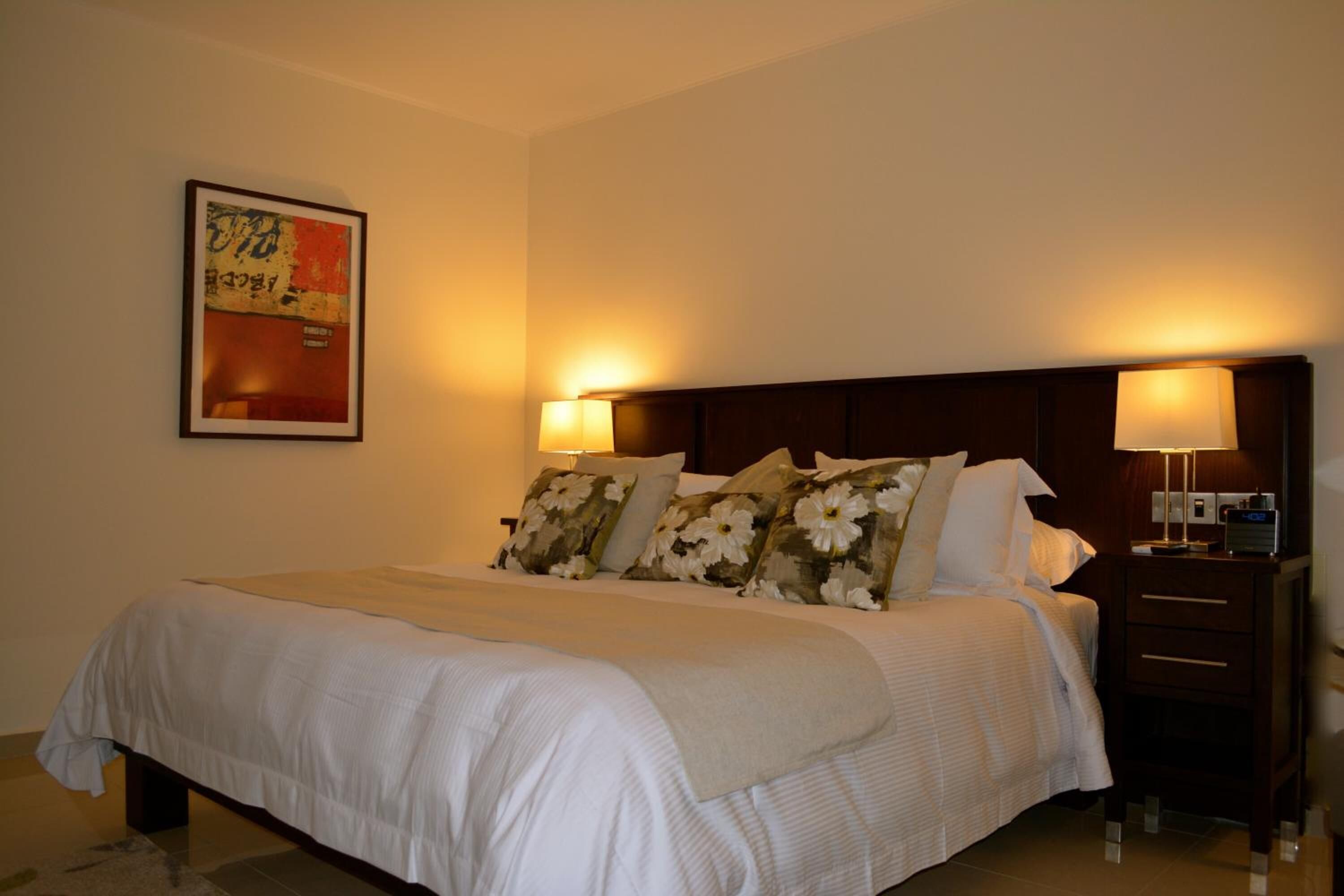 A neatly made bed with floral pillows, a bedside table with a lamp, and a framed picture on the wall.