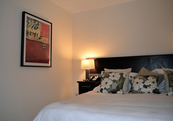 Una cama bien hecha con almohadillas florales, una lámpara de noche y una obra enmarcada en la pared.