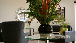Ein modernes Wohnzimmer mit einem Glastisch, einem dunklen Sessel und einer Topfpflanze mit roten Blüten.