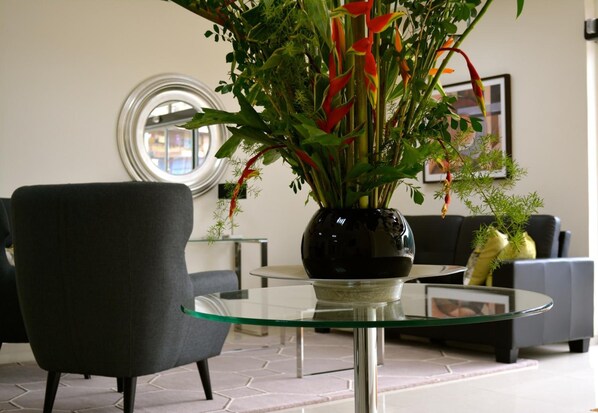 Una sala moderna con una mesa de centro de vidrio, un sillón oscuro y una planta en maceta con flores rojas.