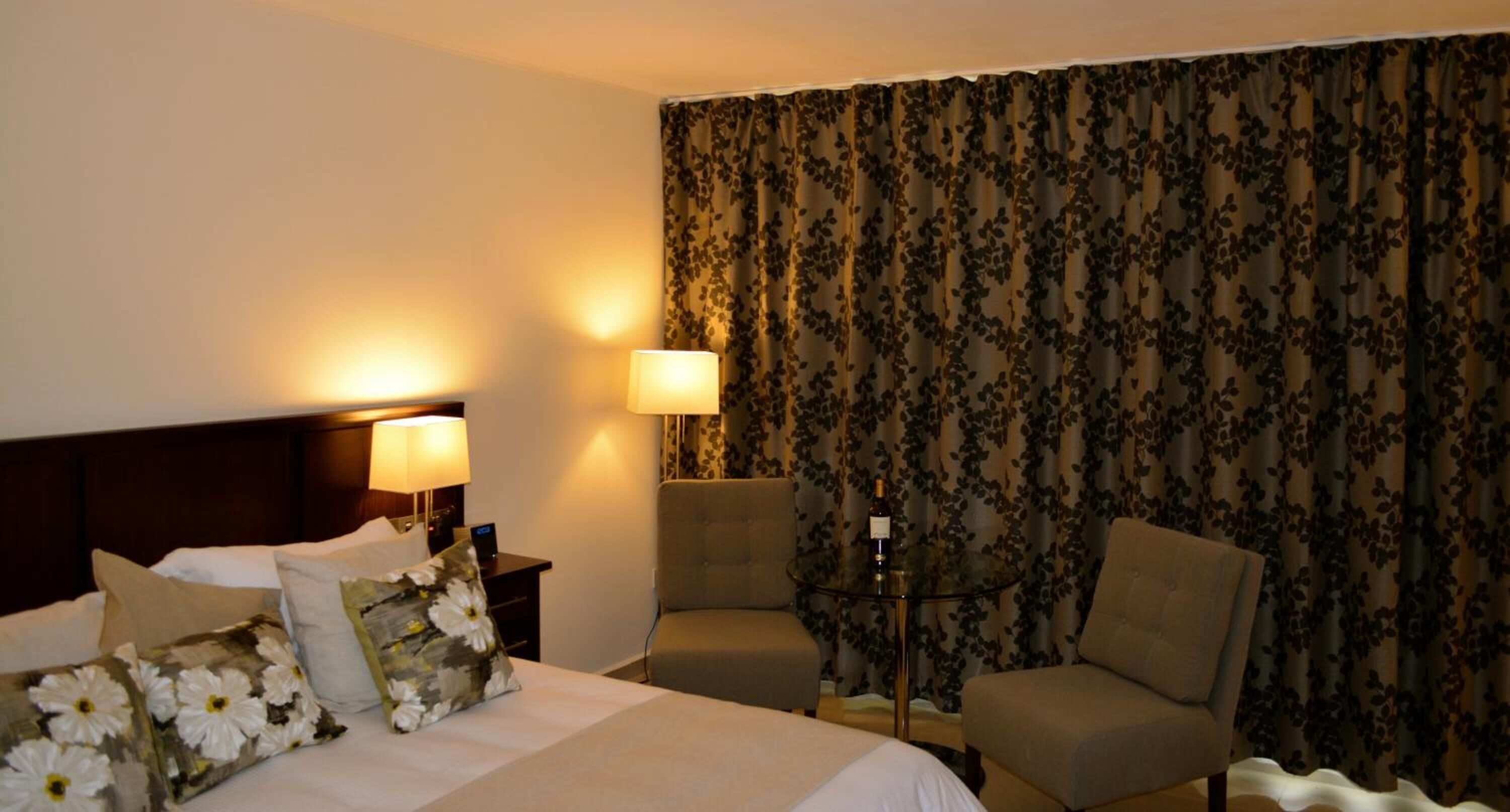 A hotel room with a bed, two chairs, a nightstand with a lamp, and a table with a bottle.