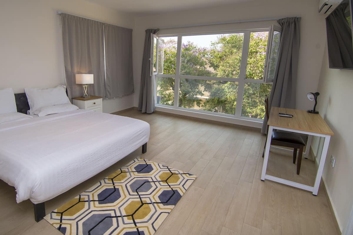 A bedroom with a large bed, a bedside table with a lamp, a small desk with a chair, and a large window with curtains.