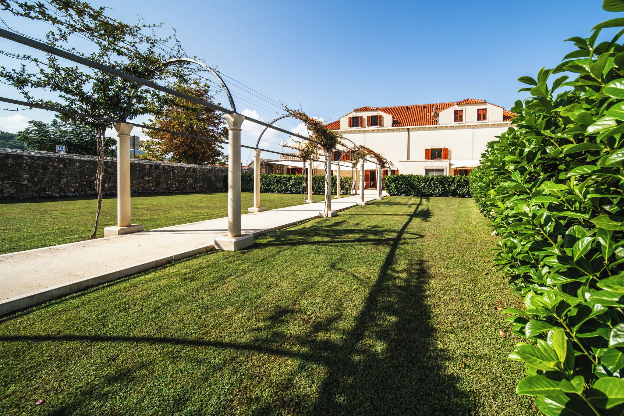 A well-maintained lawn with a pergola structure, a stone wall, and a building with a red-tiled roof.