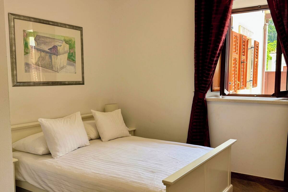 A bed with white bedding, a nightstand, a lamp, a framed picture on the wall, and a window with curtains.