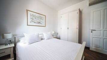 Un letto rifatto con lenzuola bianche, un comodino con una lampada, un quadro incorniciato al muro e un armadio bianco.