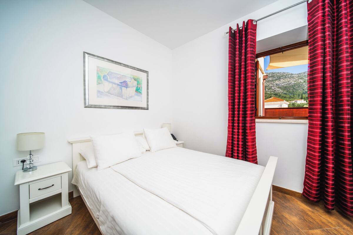 A bedroom with a bed, a nightstand, a lamp, a framed picture on the wall, and a window with red curtains.