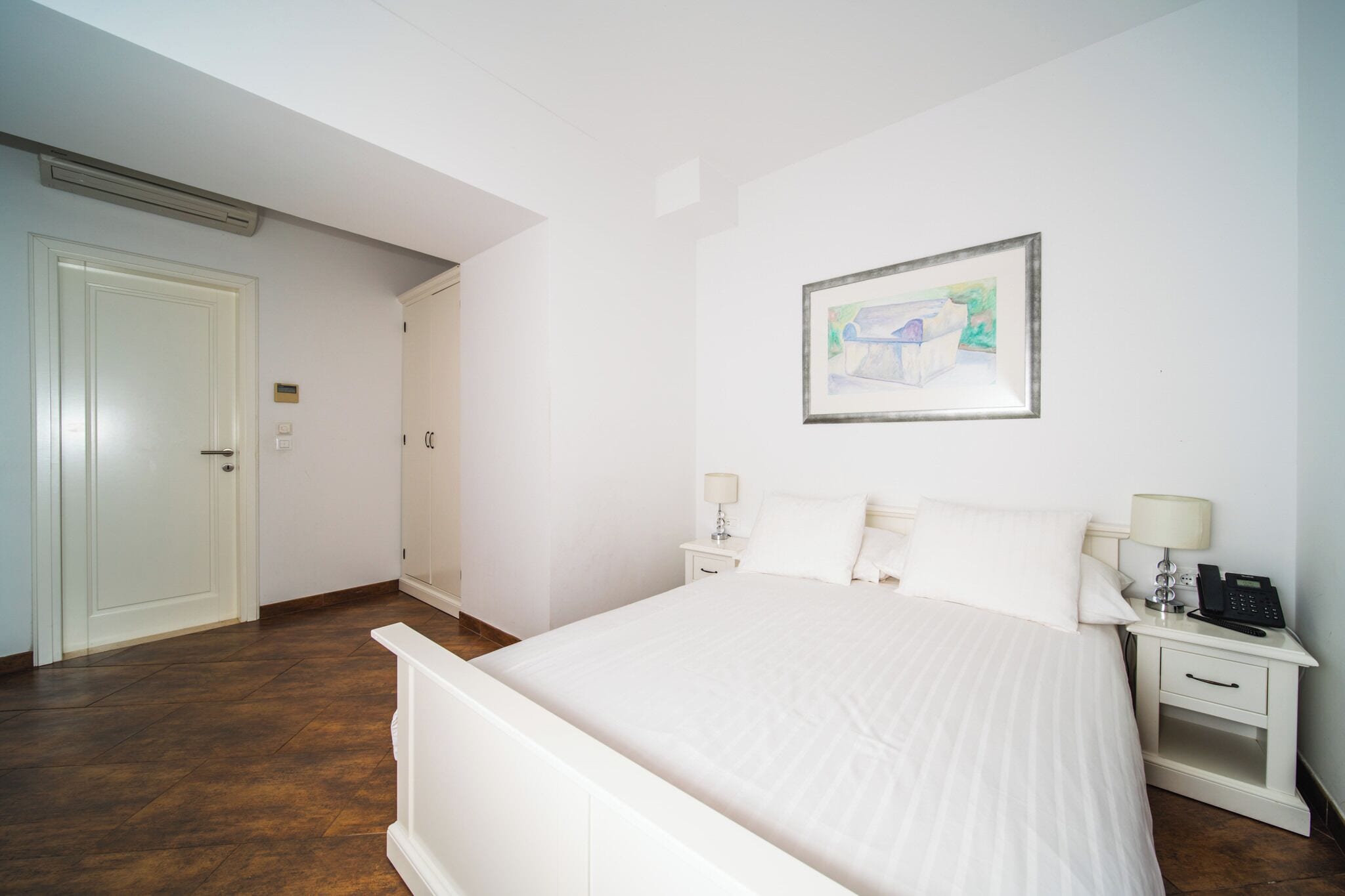 A hotel room with a large bed, white walls, a framed picture, a bedside table with a lamp, and a phone.