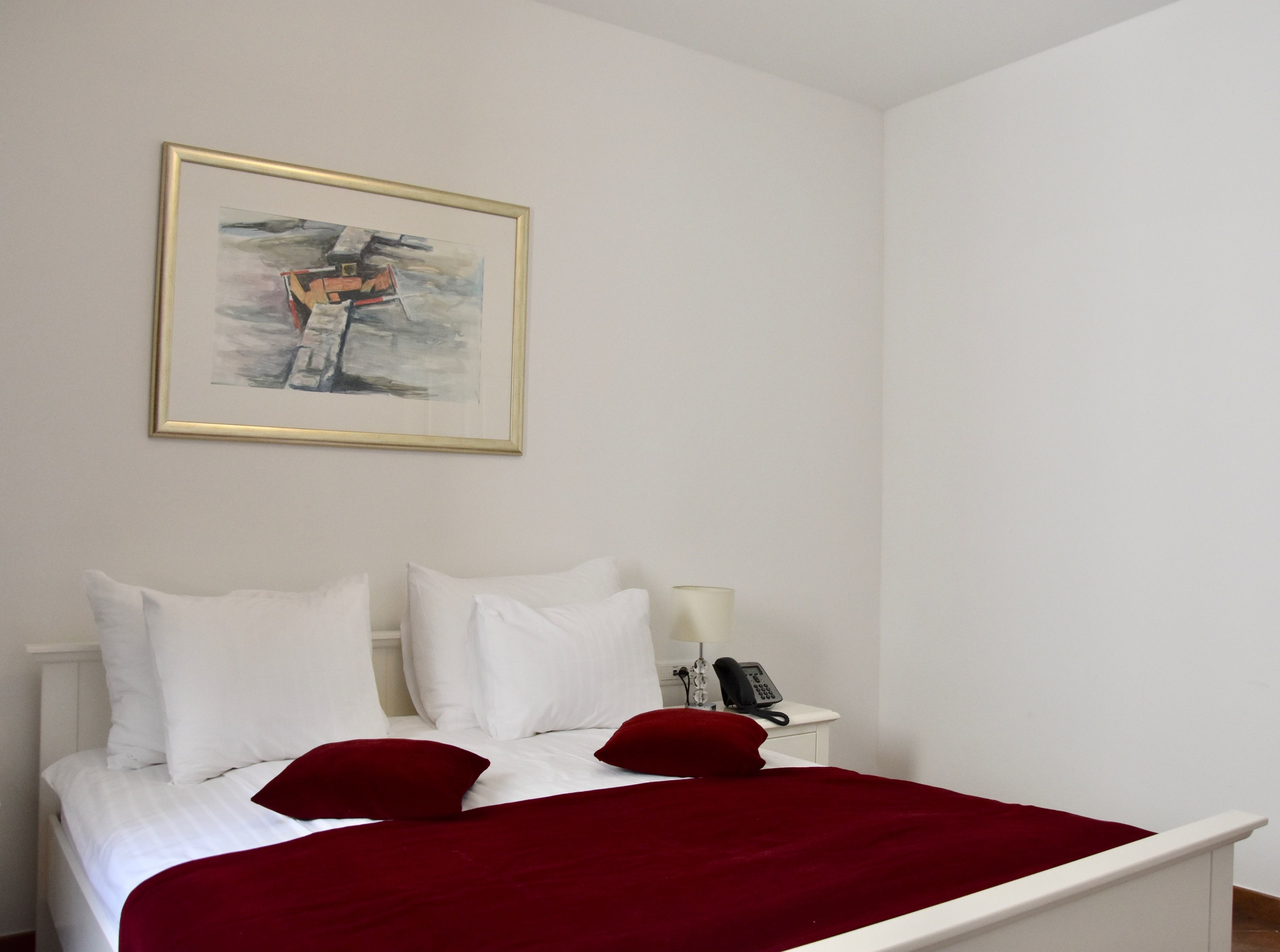 A neatly made bed with white and red bedding, a bedside phone, and a framed artwork on the wall.